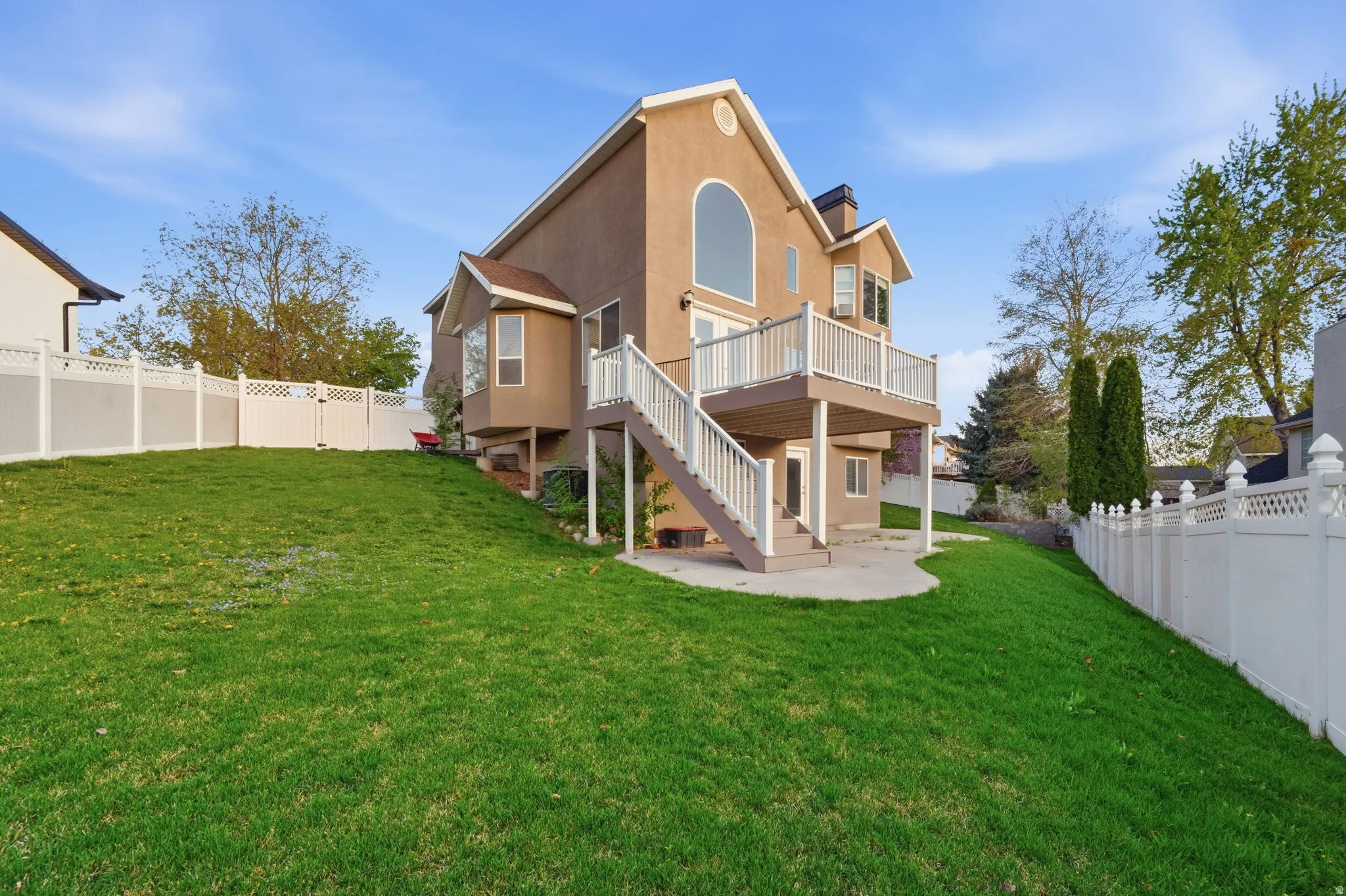 Expansive green lawn with a two-story stucco exterior, a large upper-level deck with white railings, and a walk-out lower level leading to a concrete patio