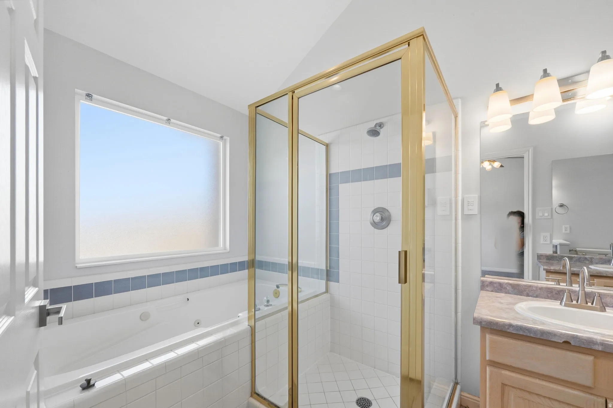 Bathroom featuring a glass-enclosed shower with a gold-tone frame, a built-in jetted tub, a single vanity with a light-toned countertop, and tile flooring
