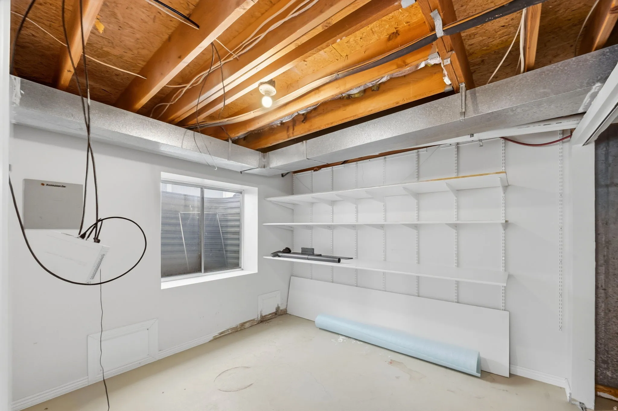 Unfinished basement space featuring exposed wood joists and ductwork, a single window, and a wall of built-in adjustable shelving