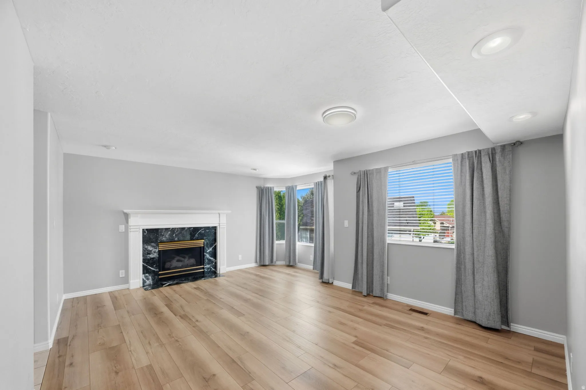 Living area featuring wood-finish flooring, a white fireplace mantel with dark stone surround, multiple windows with blinds and curtains, and recessed ceiling lighting