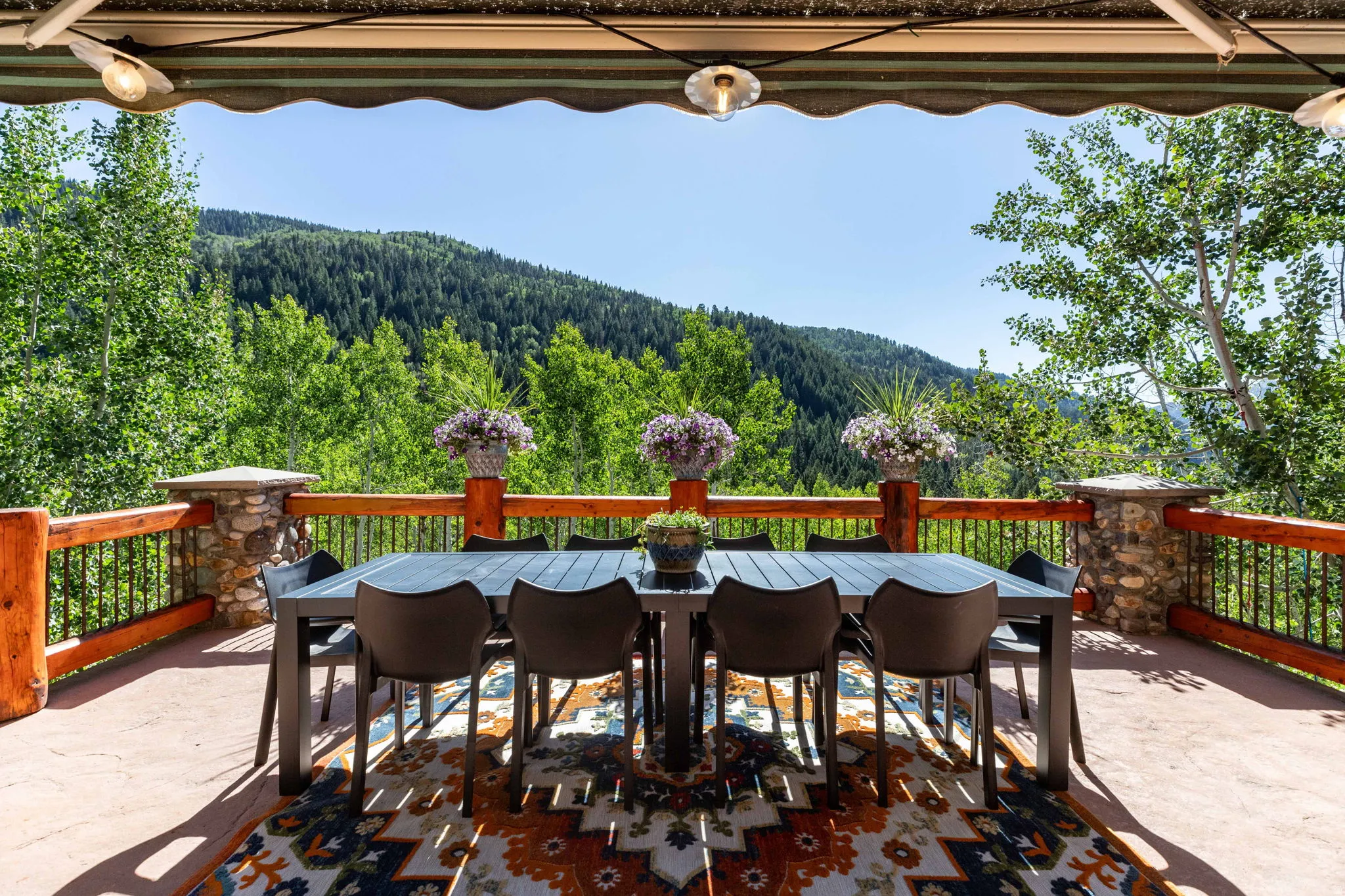 Expansive outdoor patio with a mountain backdrop, featuring a wood and wrought iron railing, stone pillars, and a retractable awning