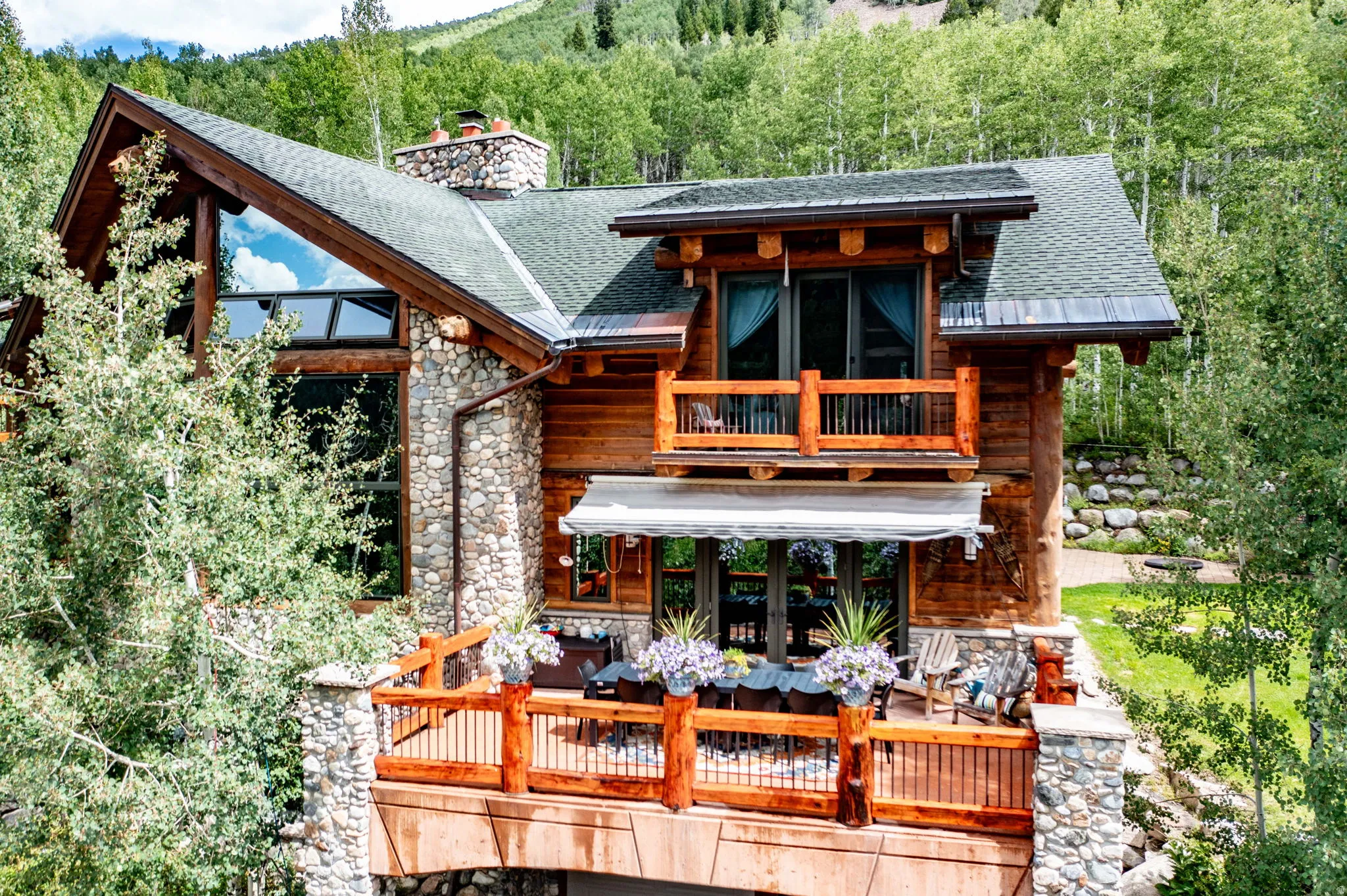 Rustic-style exterior featuring stone and log construction, a green shingle roof, a prominent stone chimney, and an expansive deck with wood railing