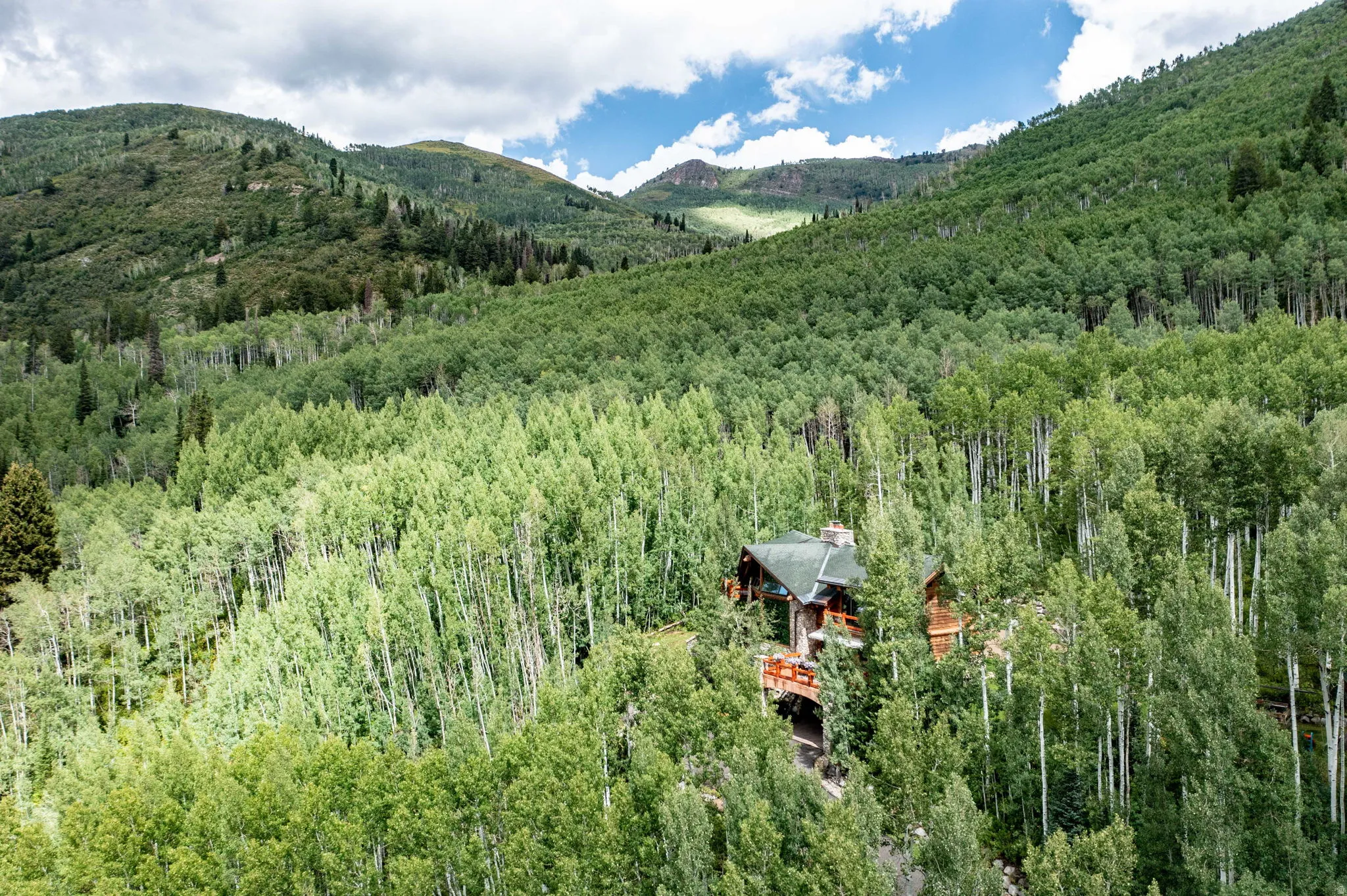 Elevated mountain home featuring a stone chimney, multiple decks, and a dark green roof, surrounded by a dense forest of deciduous trees