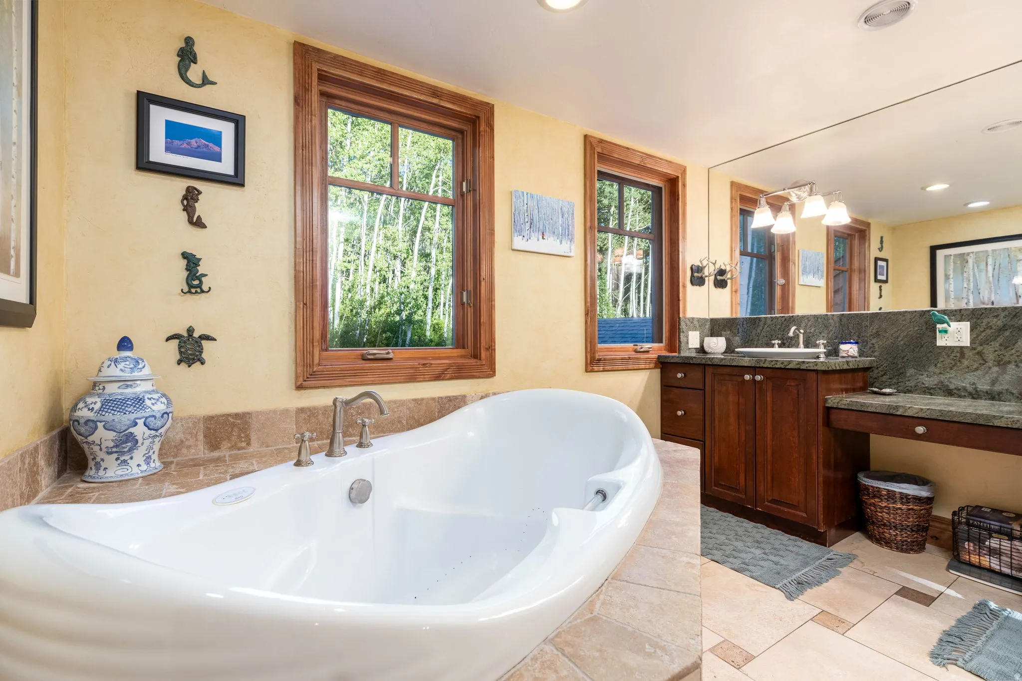 Freestanding soaking tub with brass-finish fixtures, dual wood-frame windows, stone tile flooring, and a vanity with dark wood cabinetry and a stone countertop