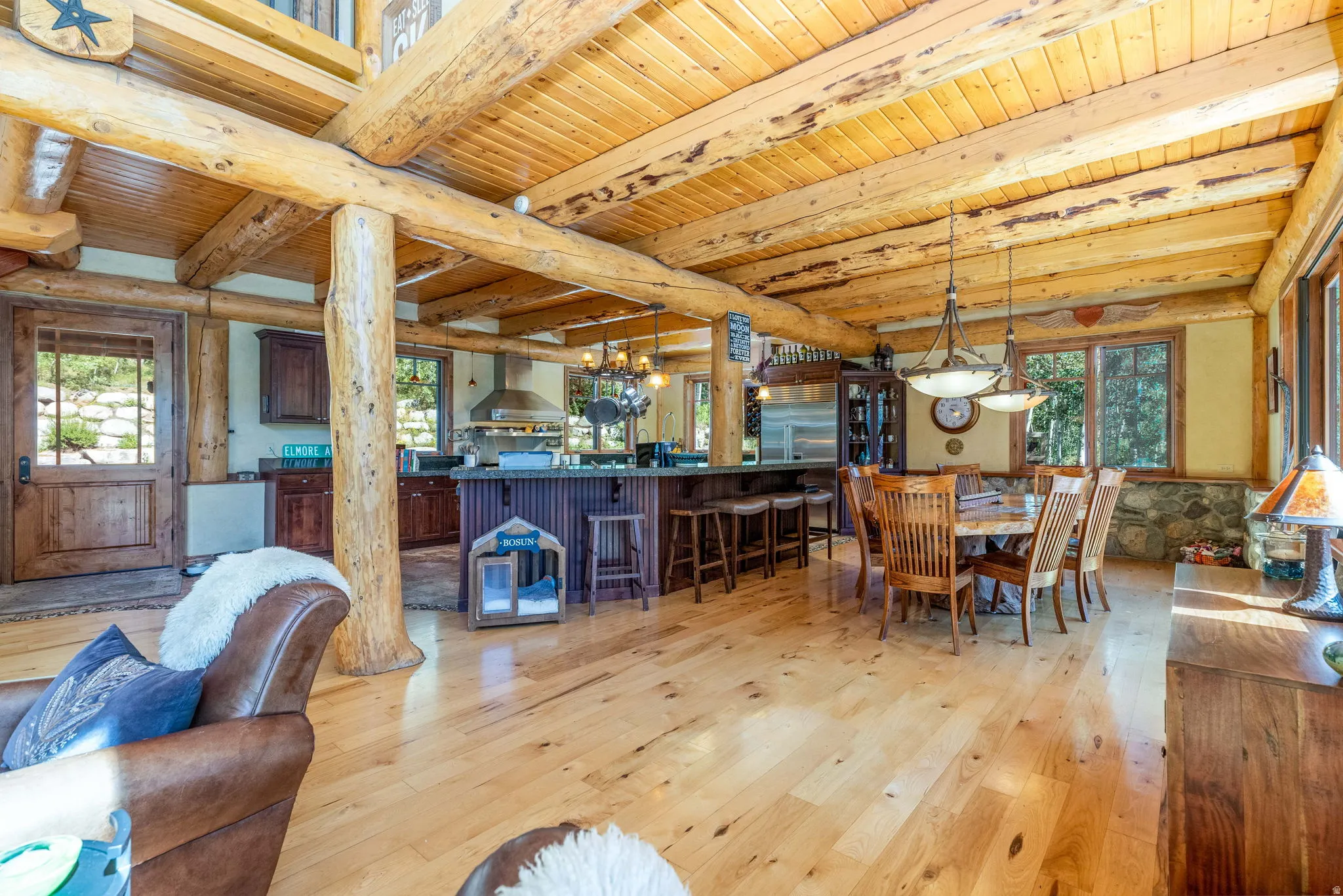 Spacious open-concept living area featuring exposed log beams, light wood-finish flooring, a kitchen island with seating, and a dining area with pendant lighting