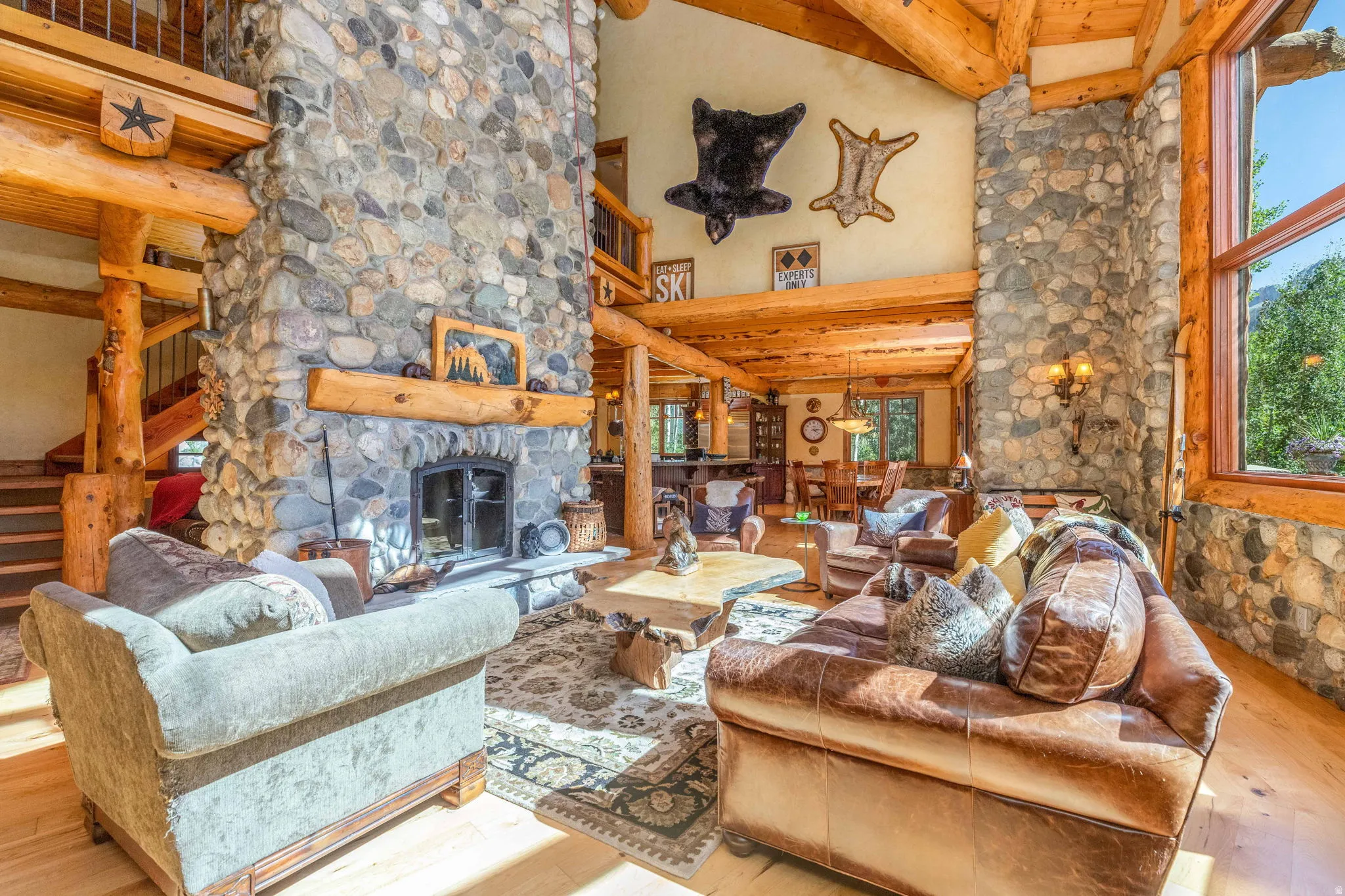 Great room featuring a floor-to-ceiling river stone fireplace with a substantial log mantel, vaulted wood-beam ceilings, expansive wood-finish flooring, a large picture window, and a visible staircase with log railings