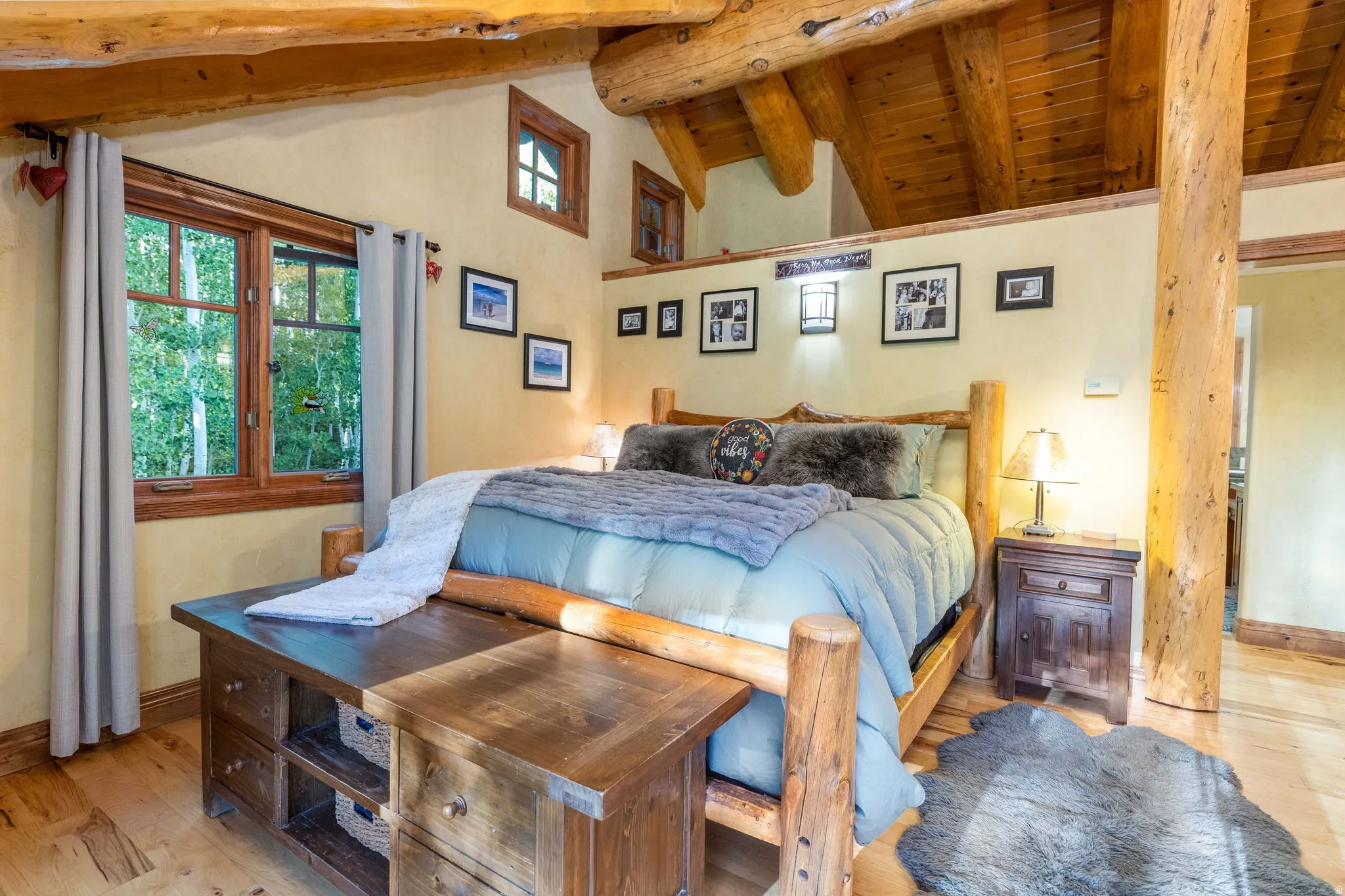 Bedroom featuring exposed log beams, wood-finish flooring, a prominent window with natural wood trim, a built-in wooden bed frame, and wall-mounted light fixtures
