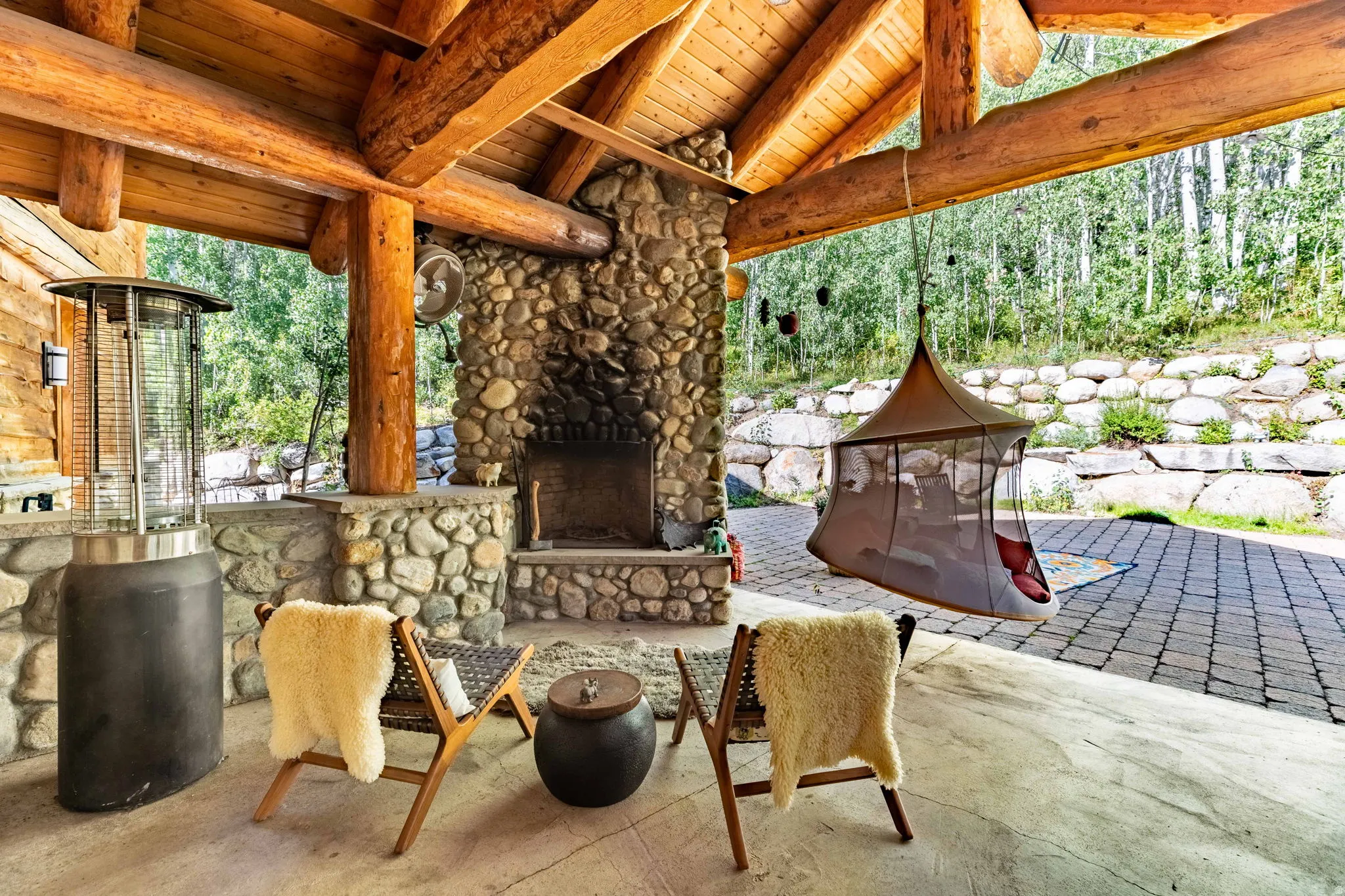 Spacious covered patio featuring exposed log beams, a stone fireplace, and a stone accent wall with built-in countertop