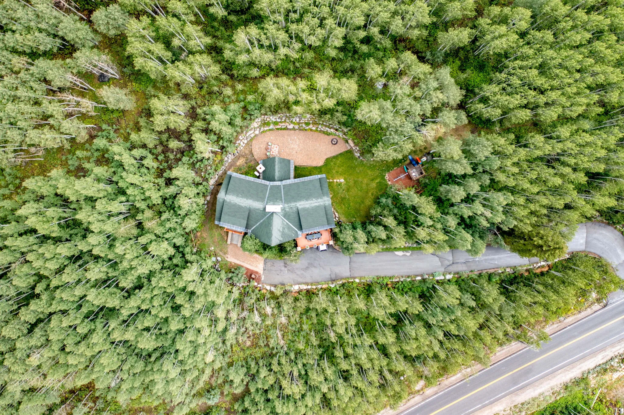 Secluded property featuring a multi-faceted green roof, surrounded by mature trees, with a stone retaining wall and a paved driveway