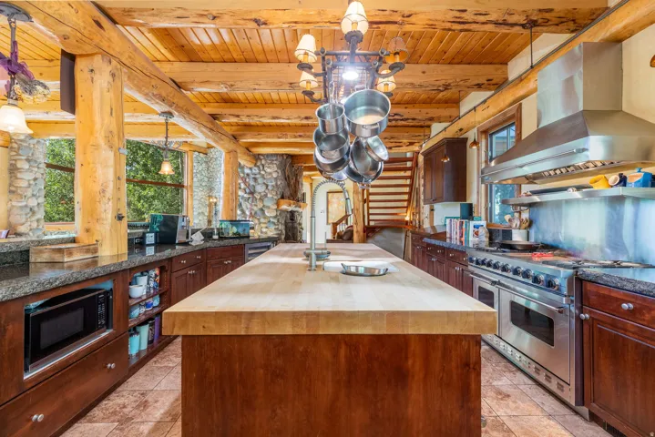 Kitchen featuring a large wood-finish island, professional-grade stainless steel range with matching hood, ample dark wood cabinetry, granite-style countertops, and exposed wood beams