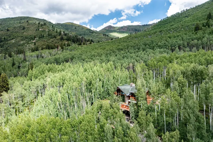 Elevated mountain home featuring a stone chimney, multiple decks, and a dark green roof, surrounded by a dense forest of deciduous trees