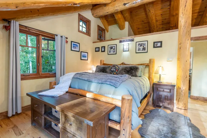 Bedroom featuring exposed log beams, wood-finish flooring, a prominent window with natural wood trim, a built-in wooden bed frame, and wall-mounted light fixtures