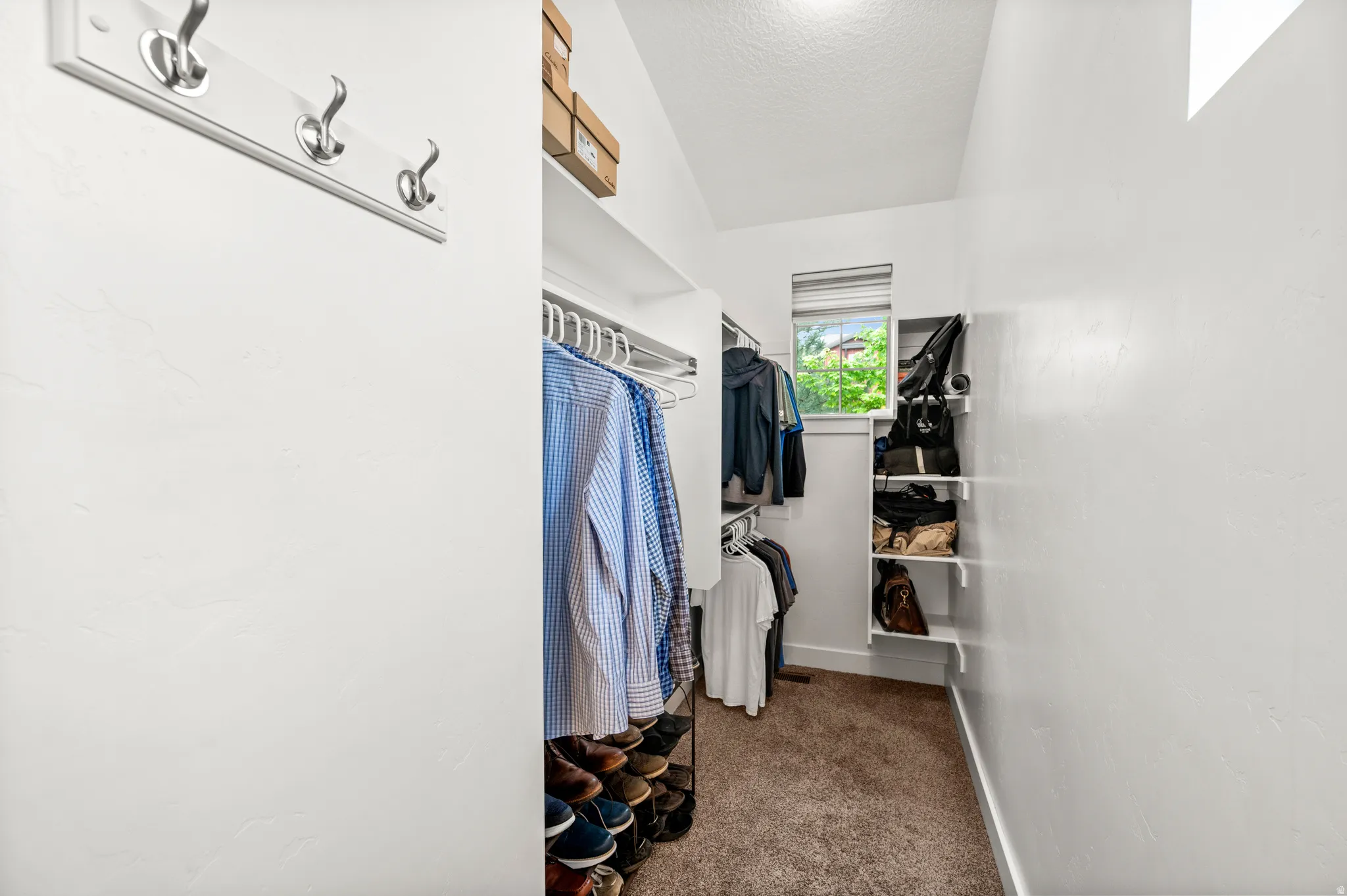 Master walk-in closet with ample hanging rods, built-in shelving, carpet flooring, a window providing natural light, and a wall-mounted coat rack