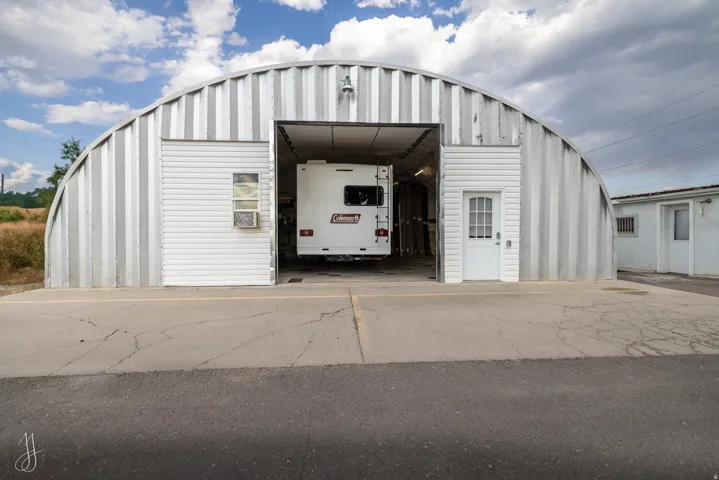 Quonset hut-style metal building featuring a large overhead door, white vinyl siding accents, a glass panel pedestrian door, and a window with an air conditioning unit