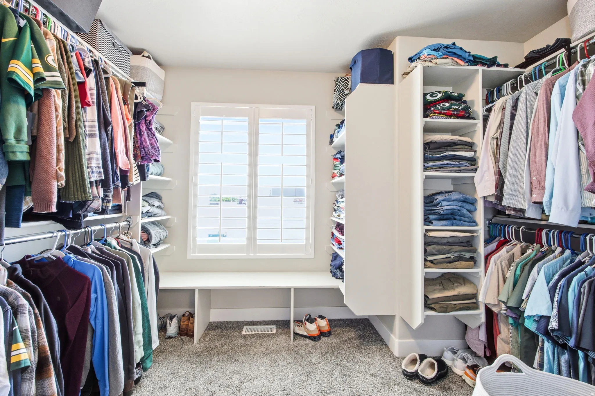 Walk-in closet featuring custom built-in shelving, hanging rods, an integrated bench, and window with plantation shutters