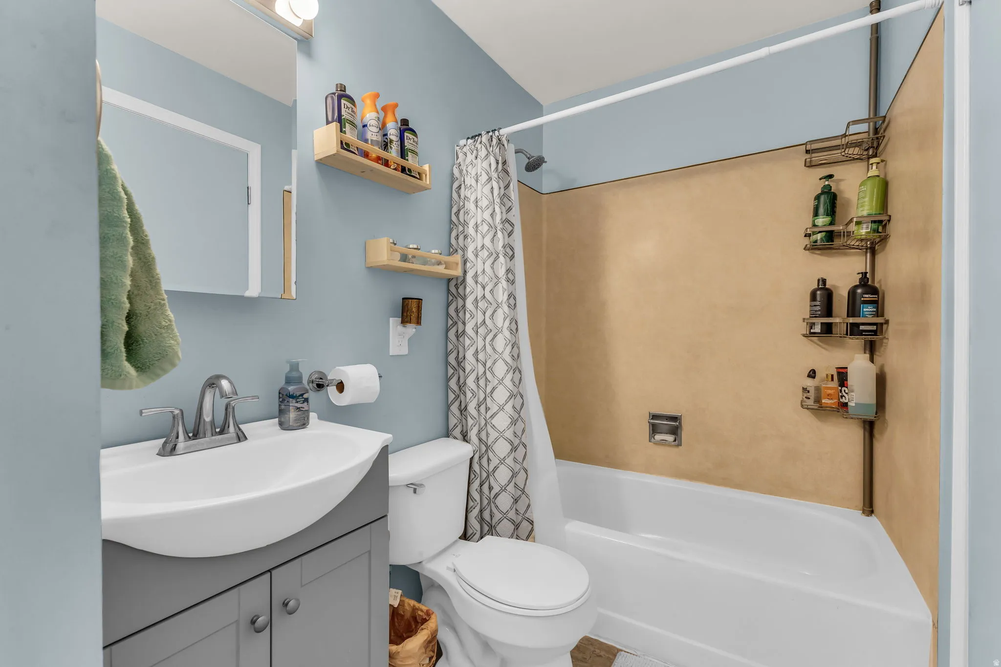 Bathroom featuring a white porcelain toilet, a shower-over-tub combination with a light brown surround, and a vanity with a white ceramic sink and gray cabinetry