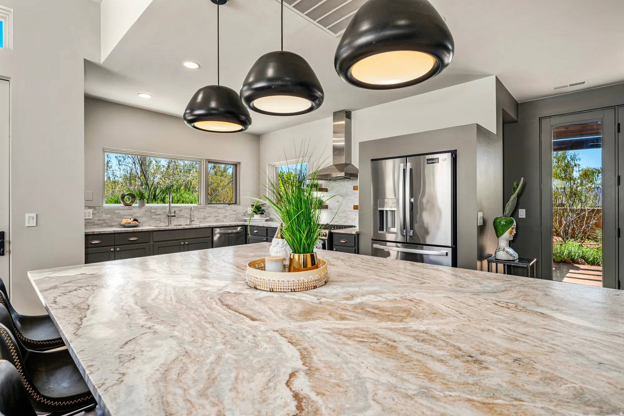 Spacious kitchen island featuring a natural stone countertop, three pendant lights, stainless steel appliances, dark cabinetry, and a tile backsplash