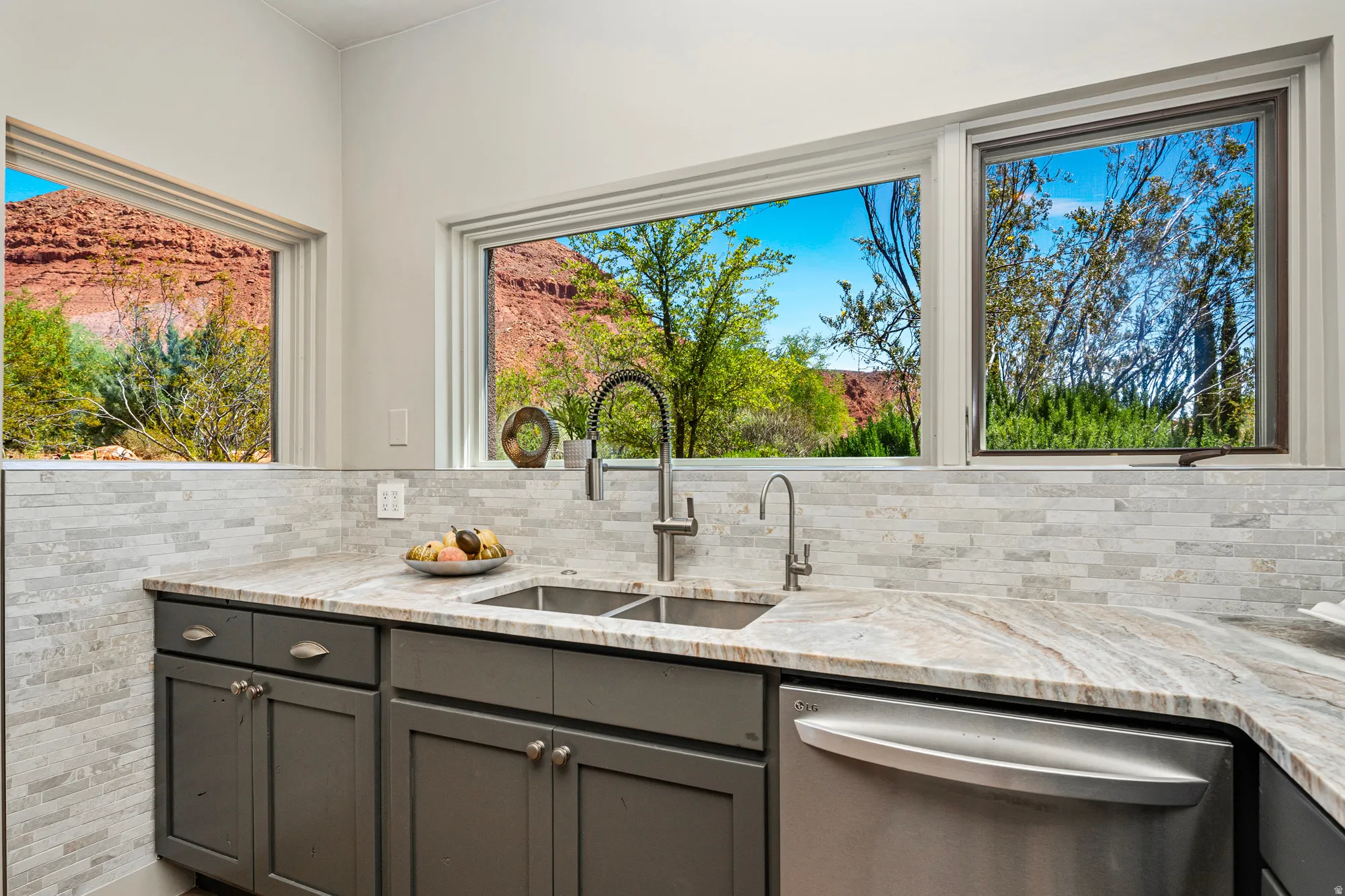 Kitchen featuring dual basin stainless steel sink with gooseneck faucet, light-toned stone countertops, and gray shaker-style cabinetry with matte hardware