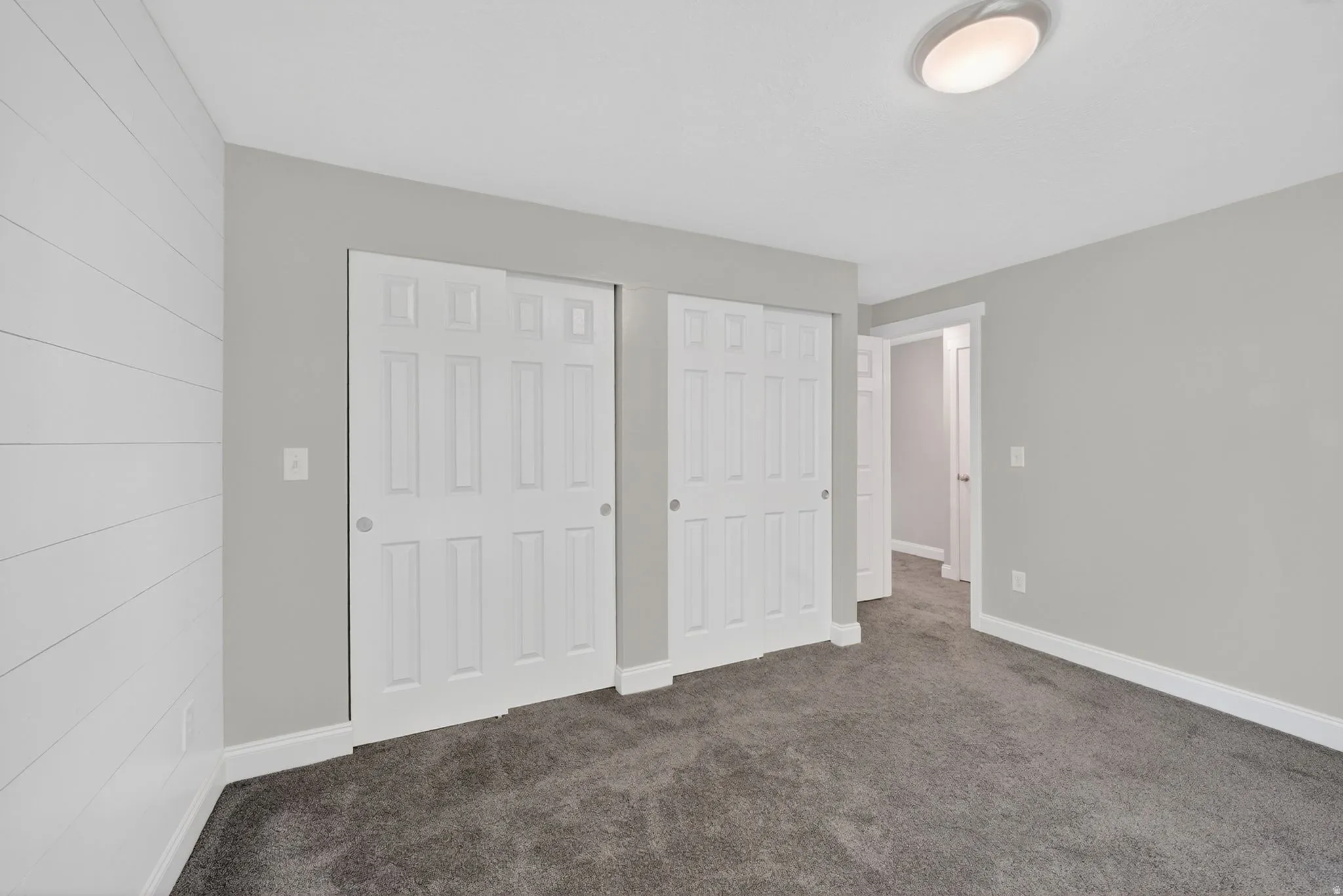 Room featuring a shiplap accent wall, two bifold closet doors, light gray wall paint, dark gray carpeting, and a flush-mount ceiling light