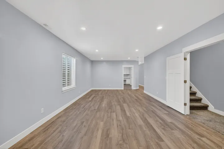 Spacious room featuring wood-finish flooring, light blue walls, recessed ceiling lighting, a window with blinds, and white trim