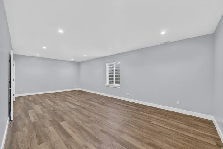 Spacious room featuring wood-finish flooring, light blue walls, recessed lighting, and a window with white louvered blinds