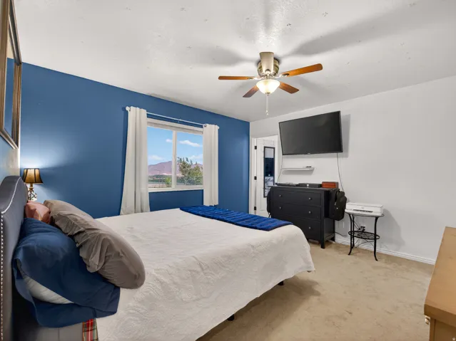 Primary bedroom featuring a window with distant mountain scenery, neutral carpeting, a ceiling fan with integrated lighting, and a wall-mounted television