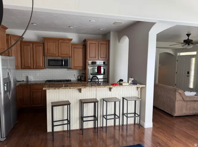 Kitchen featuring wood-finish flooring, warm wood cabinetry, stainless steel appliances, a white subway tile backsplash, and a breakfast bar with beadboard paneling