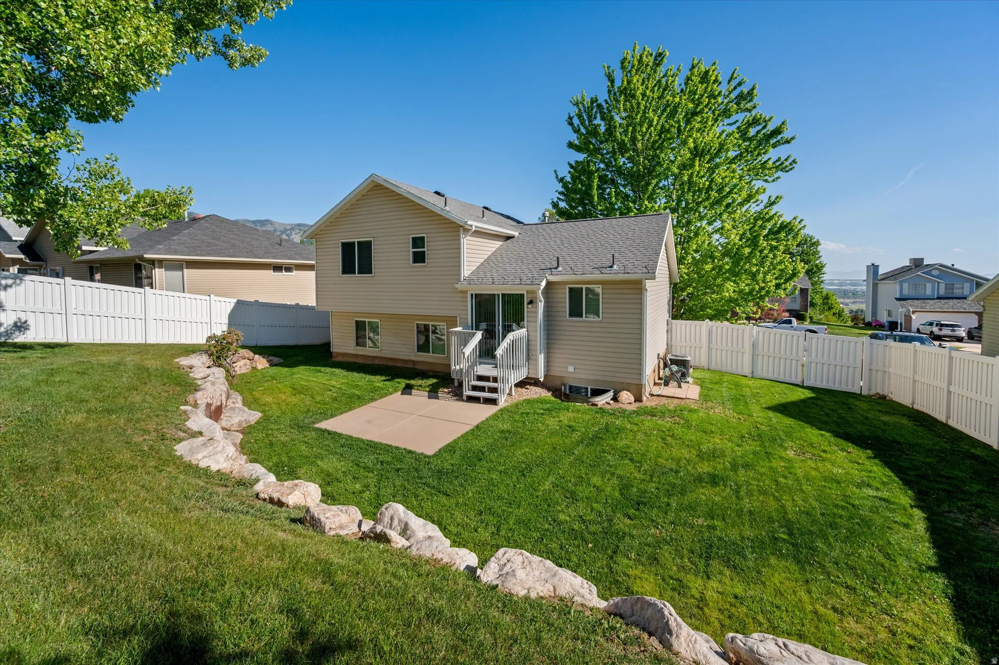 Expansive grassy yard featuring a multi-level house exterior with siding, a white privacy fence, a concrete patio, and a deck with stairs