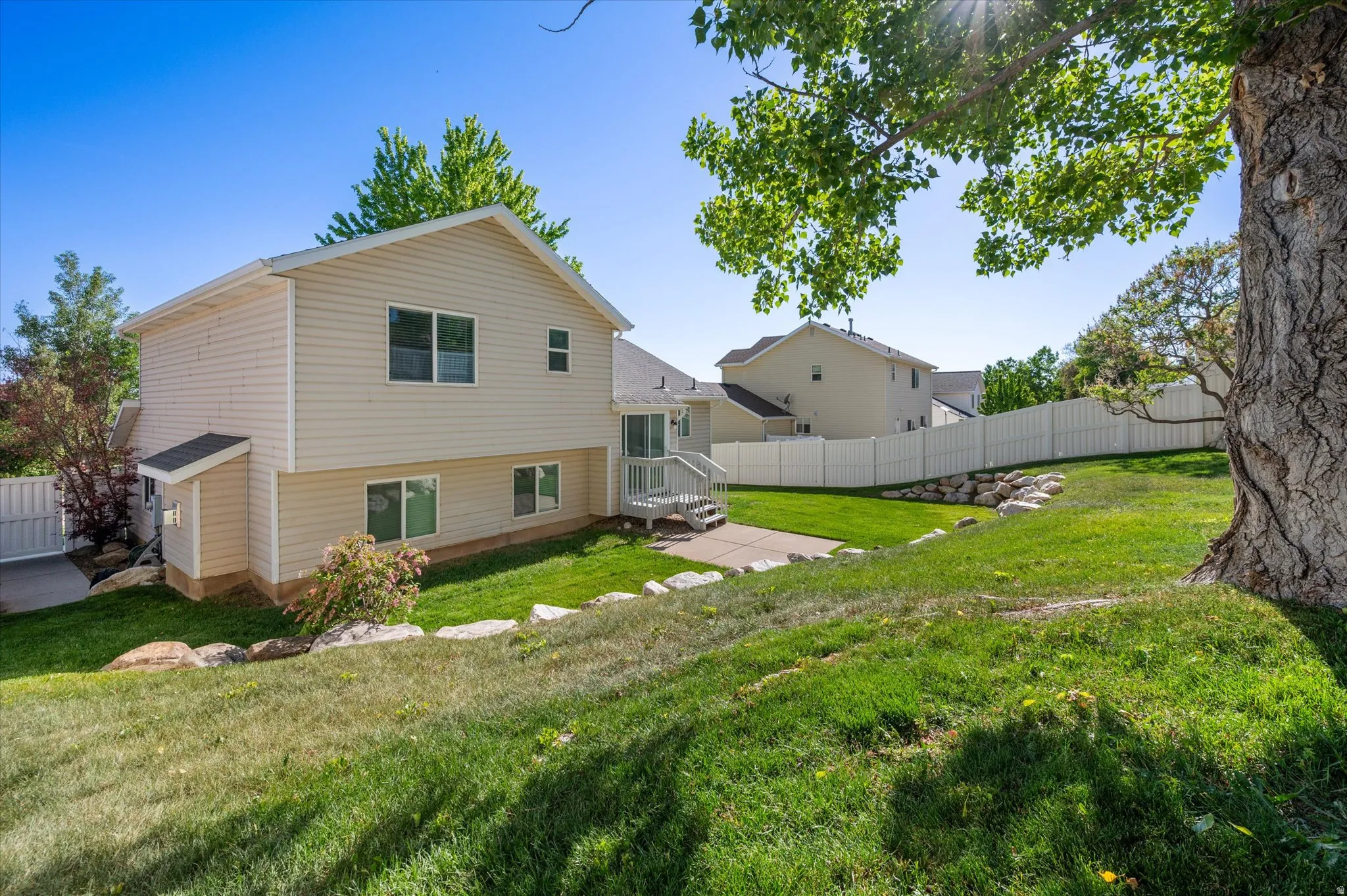 Expansive sloped backyard featuring a manicured lawn, established shade tree, and a concrete patio