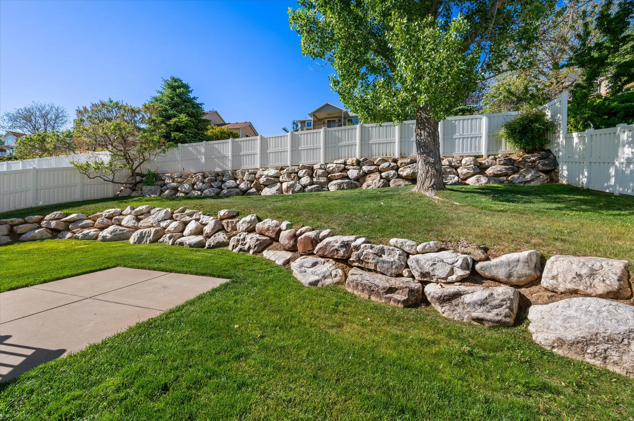 Expansive backyard featuring a concrete patio, verdant lawn, multi-tiered rock retaining walls, mature trees, and privacy fencing