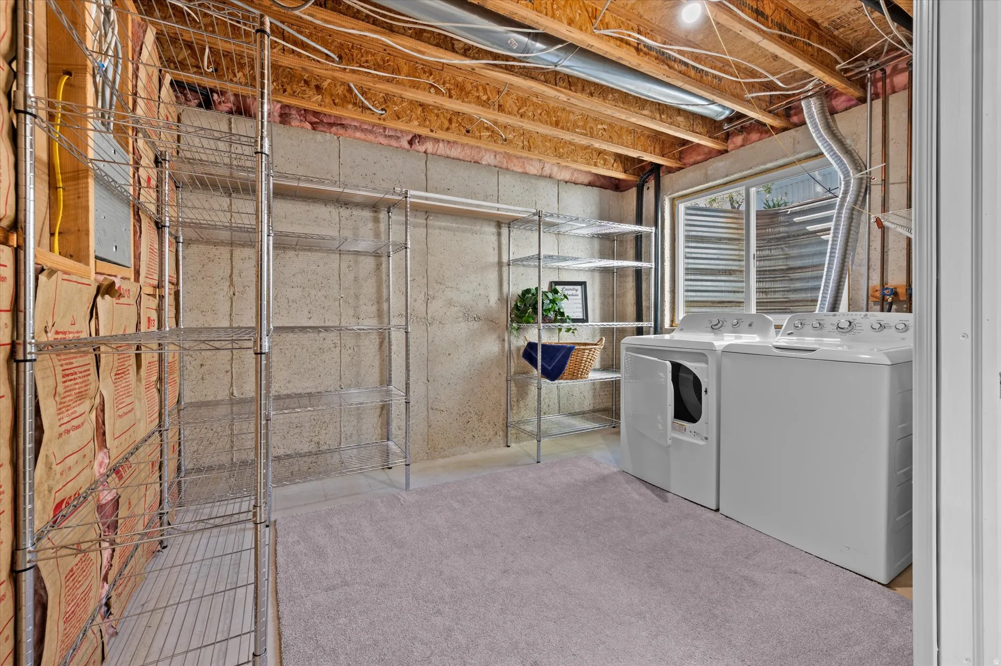 Laundry area featuring a window, bare concrete walls, exposed ceiling joists, and light gray carpeting