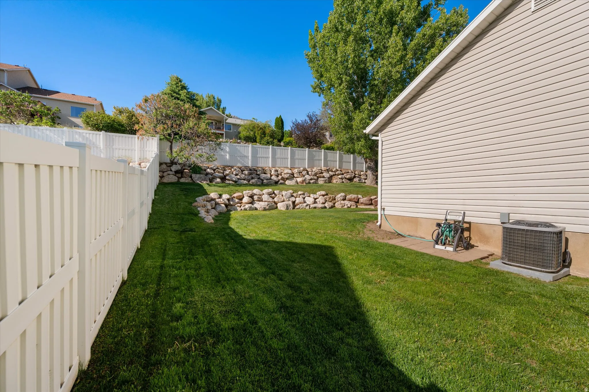 Expansive backyard featuring a manicured lawn, white privacy fencing, and a rock retaining wall