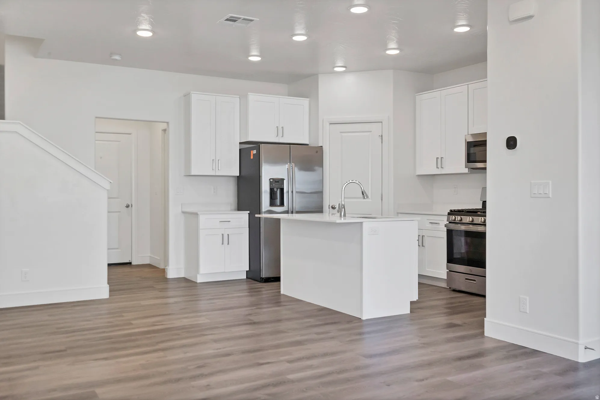 Open-concept kitchen featuring white cabinetry, stainless steel appliances, a central island with sink, and wood-finish flooring