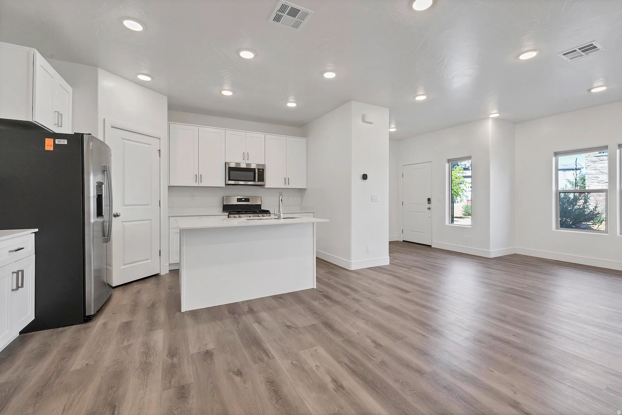 Open-concept living area featuring wood-finish flooring, recessed lighting, and a kitchen with white cabinetry, stainless steel appliances, and a central island with a sink