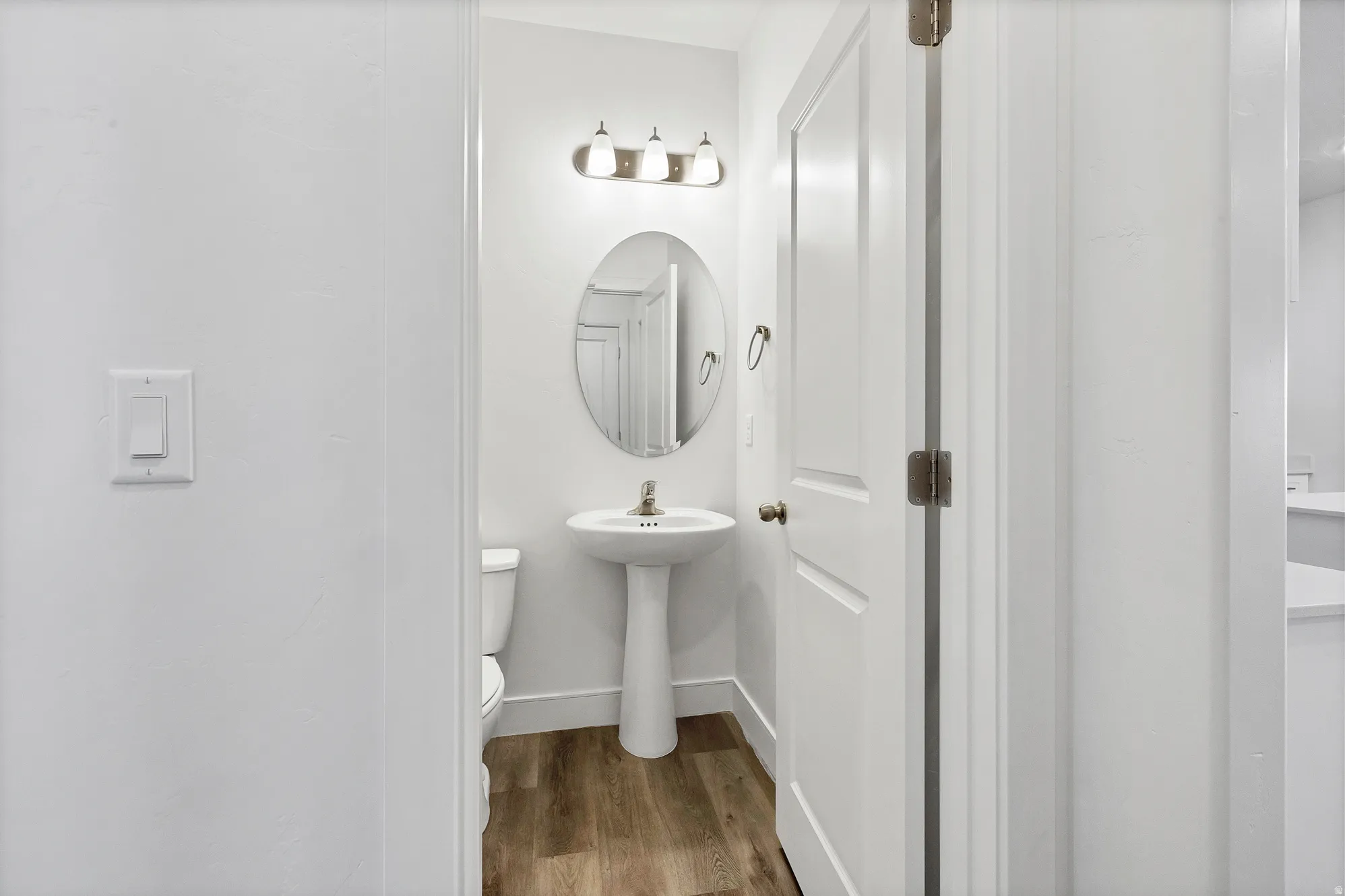 Compact bathroom featuring a white pedestal sink, oval mirror, and wood-finish flooring
