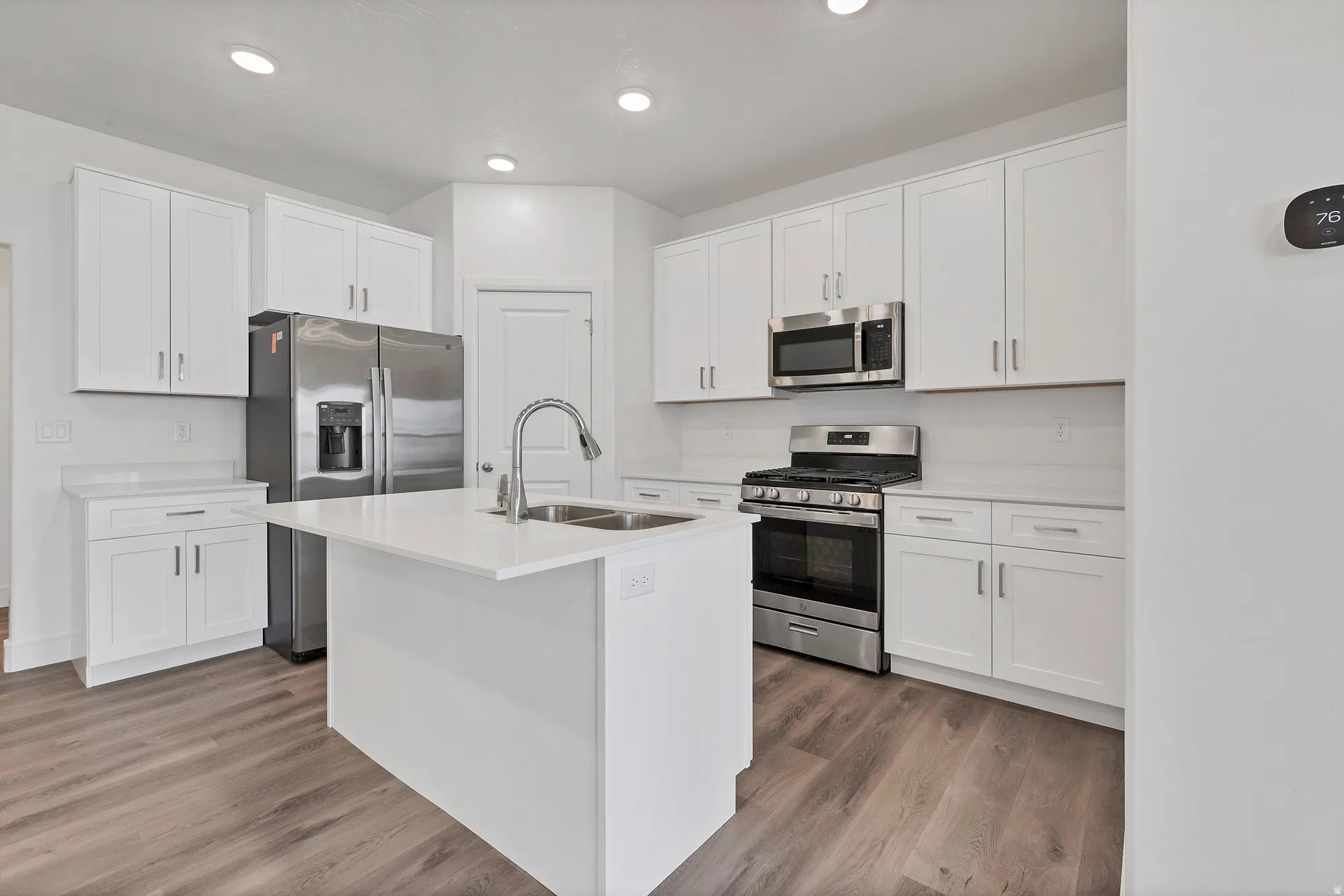 Contemporary kitchen featuring a central island with an undermount sink, white cabinetry with brushed nickel hardware, light-toned countertops, wood-finish flooring, and stainless steel appliances