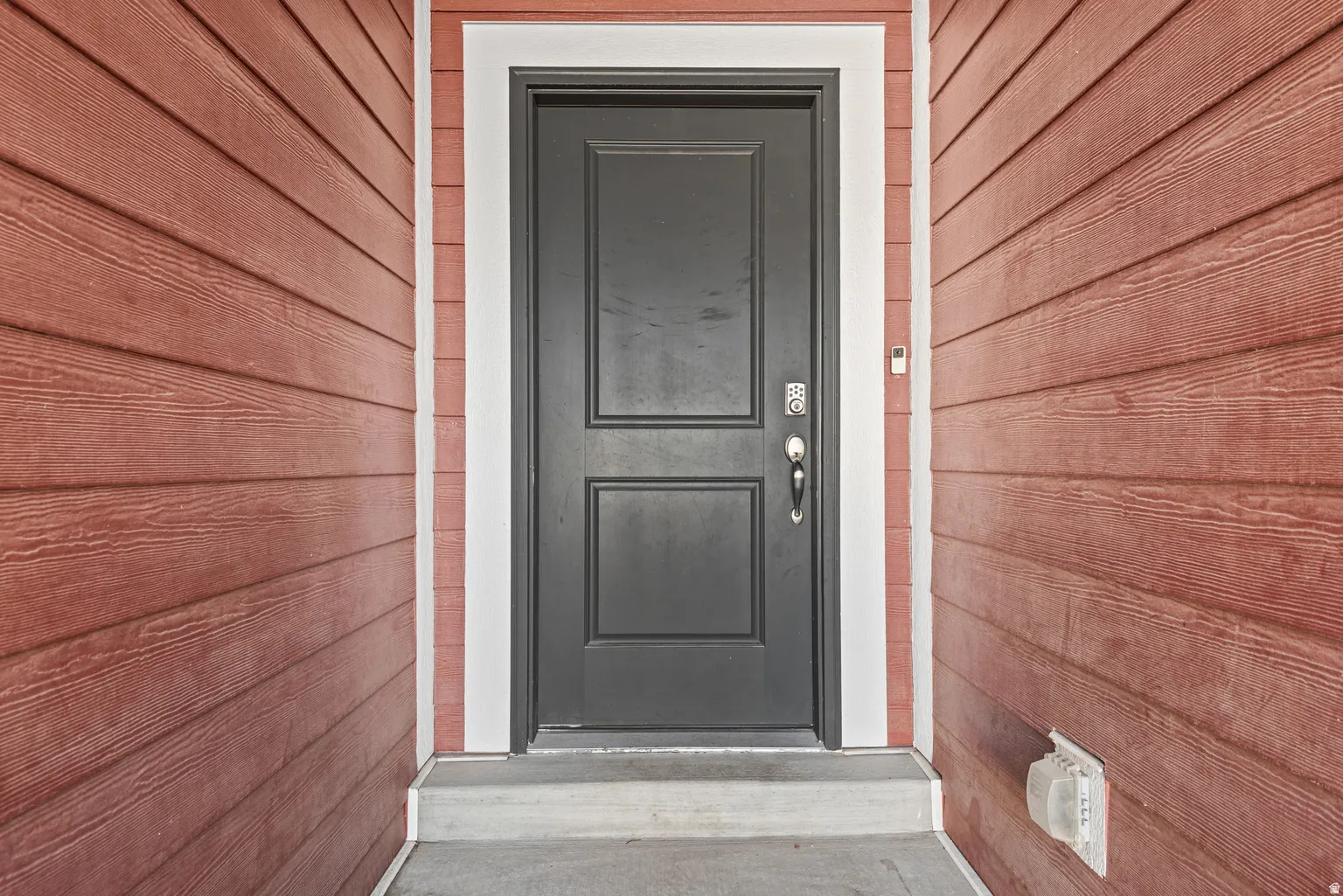 Dark paneled entry door with keypad entry, framed by white trim and red wood-finish siding