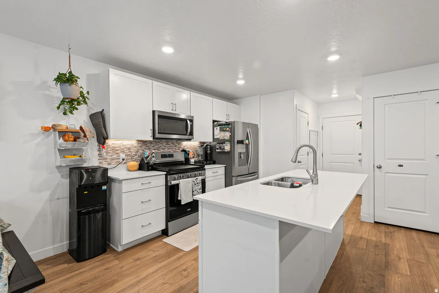 Modern kitchen featuring an island with an undermount sink, white cabinetry, stainless steel appliances, a mosaic tile backsplash, and wood-finish flooring