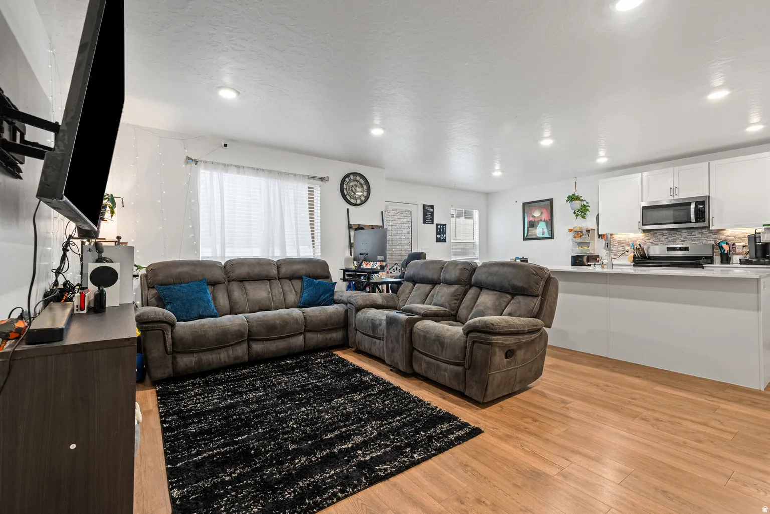 Open-concept living area featuring wood-finish flooring, recessed lighting, and a kitchen with white cabinetry, stainless steel appliances, and a tiled backsplash