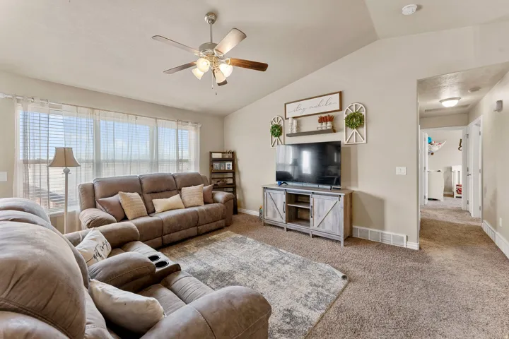 Living area featuring vaulted ceilings, a ceiling fan, a large window with horizontal blinds, and carpet flooring