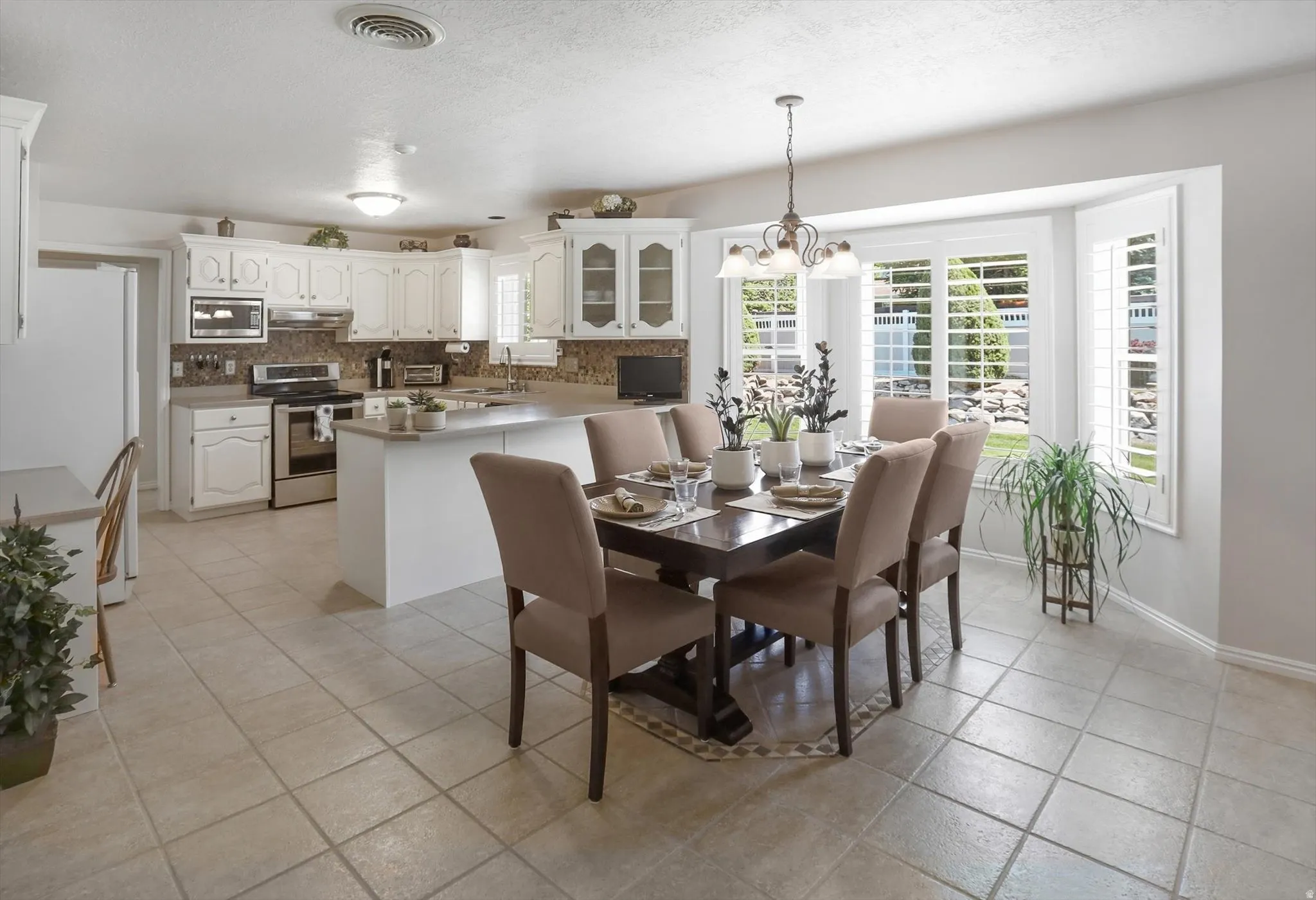 Open-concept kitchen and dining area featuring white cabinetry, stainless steel appliances, a bay window with plantation shutters, tile flooring, and a dining table with seating