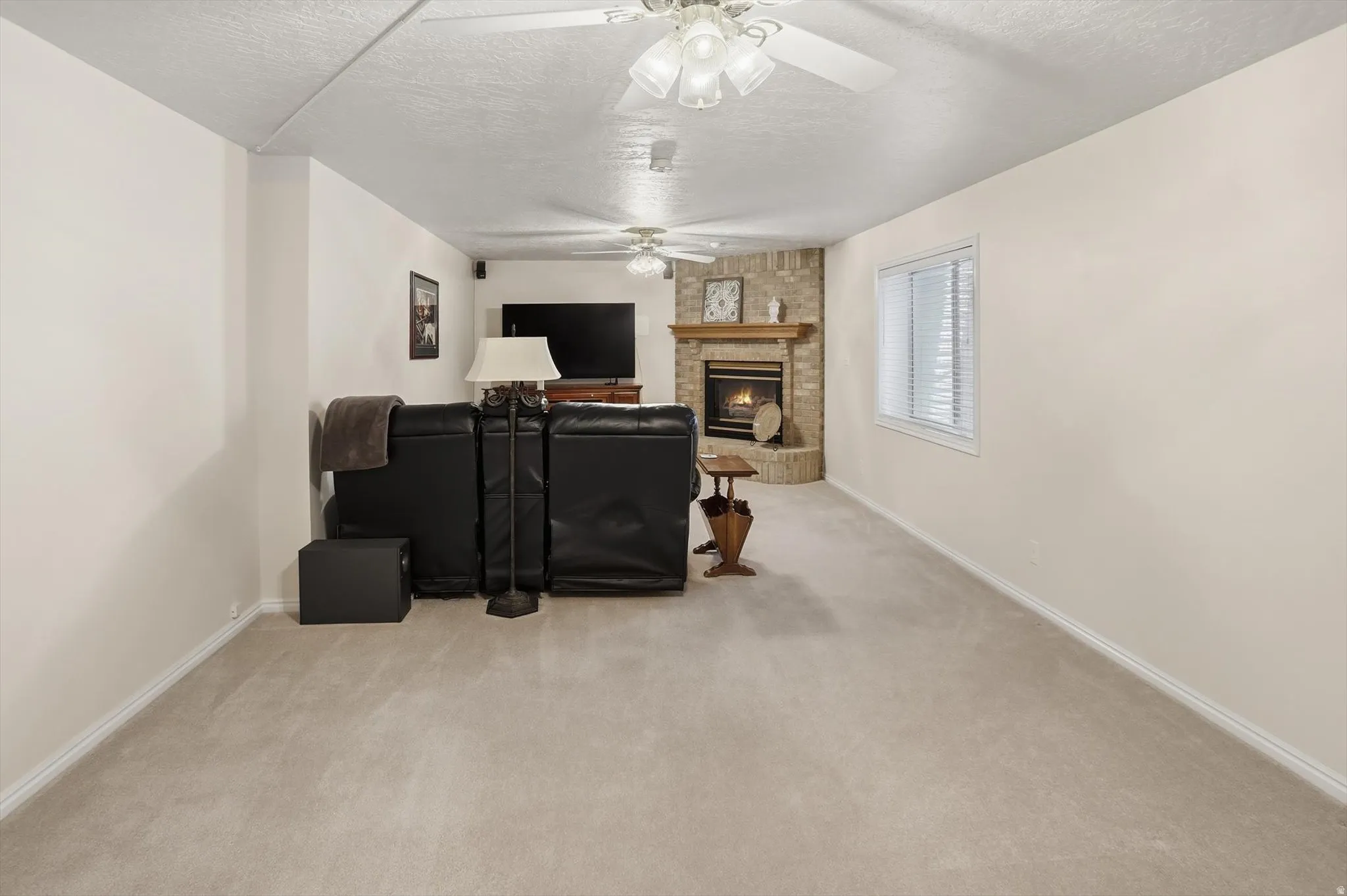 Spacious living area featuring neutral carpeting, a corner brick fireplace with mantel, white walls, and built-in ceiling fans