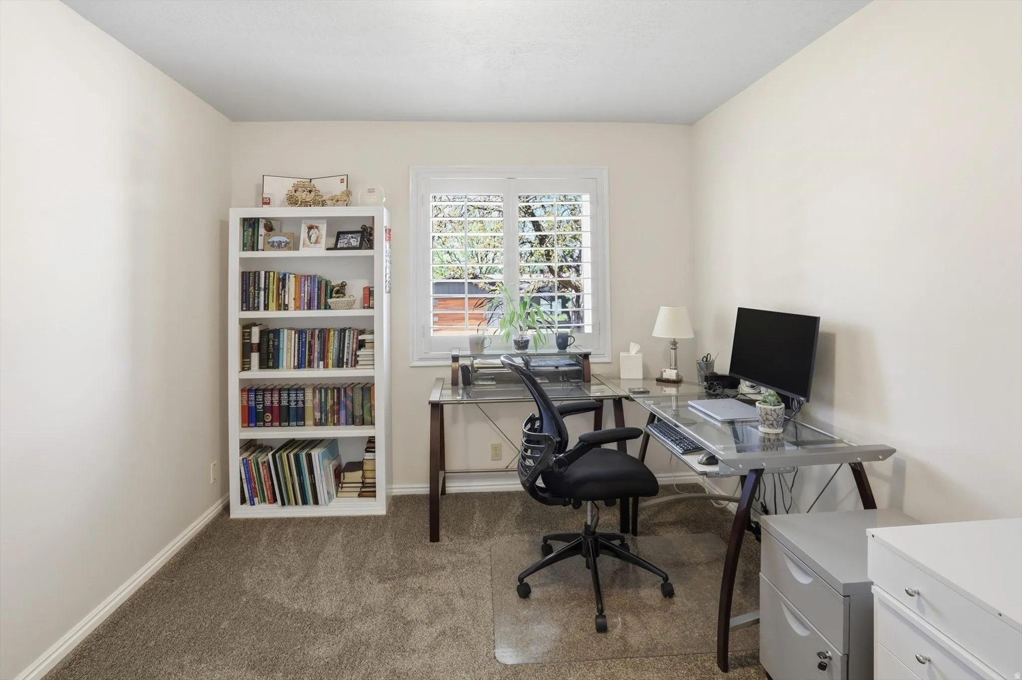 Light-colored walls with a window featuring white plantation shutters, complemented by a built-in white bookshelf and neutral carpet flooring