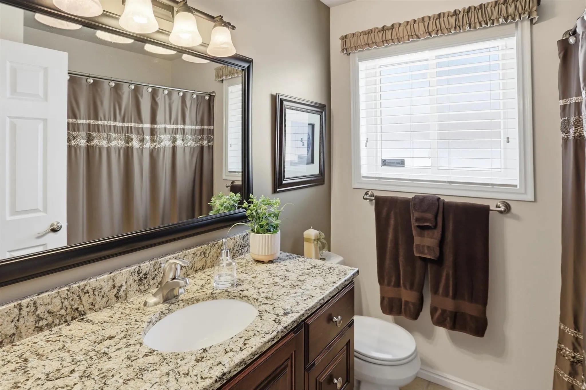 Bathroom featuring a granite countertop vanity with an undermount sink, dark wood cabinetry, a large framed mirror, and a window with blinds