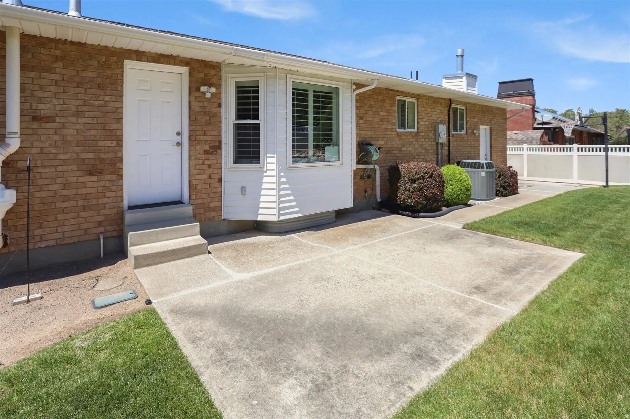 Rear exterior featuring a concrete patio, a bay window with plantation shutters, brick siding, and a white vinyl fence