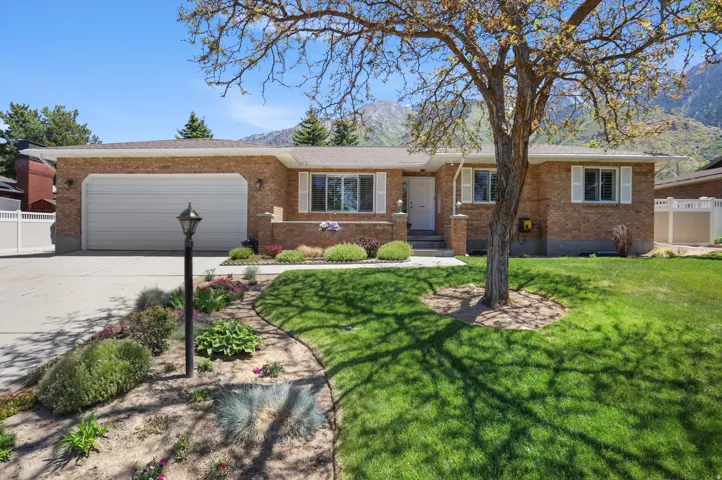 Brick exterior residence featuring a two-car garage, mature front yard tree, established landscaping, and a mountain backdrop
