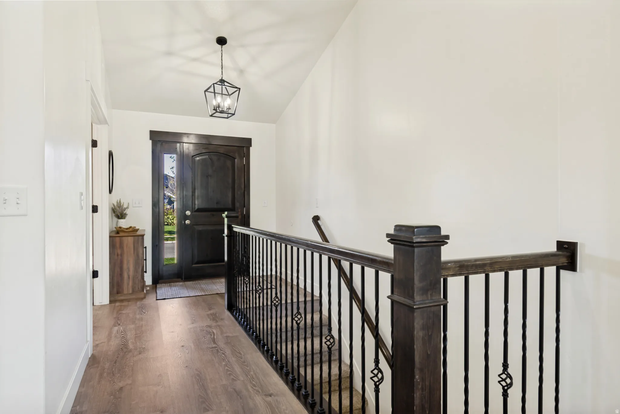Entryway featuring a dark wood-finish door with sidelights, wood-finish flooring, a vaulted ceiling, a geometric pendant light fixture, and a dark wood-finish staircase railing with wrought iron balusters.