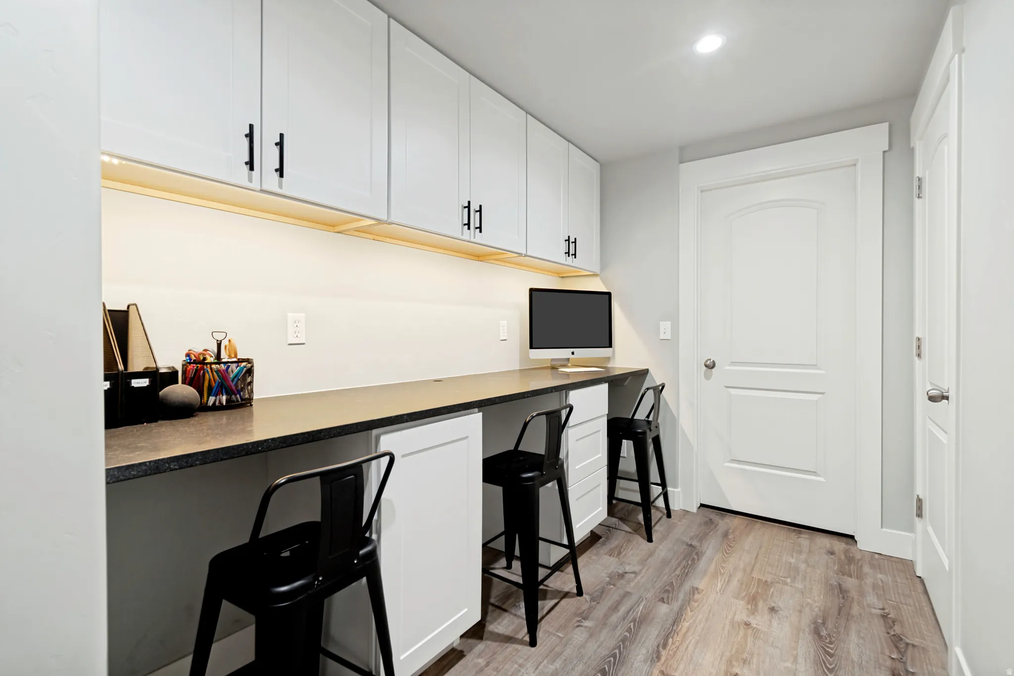 Dedicated workspace featuring light wood-finish flooring, an extended countertop, under-cabinet lighting, multiple white upper cabinets with matte black hardware, and two white lower cabinets.