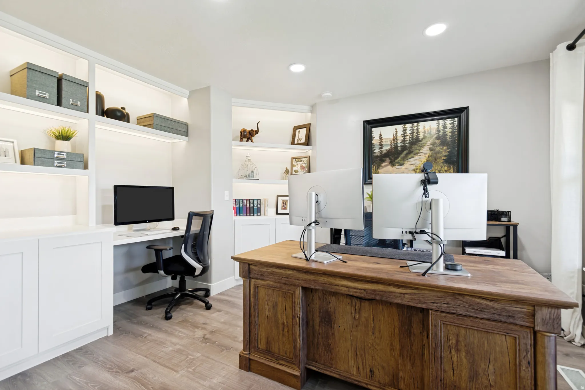 Built-in shelving with integrated lighting, wood-finish flooring, recessed ceiling lighting, a dedicated desk area, and a large window.