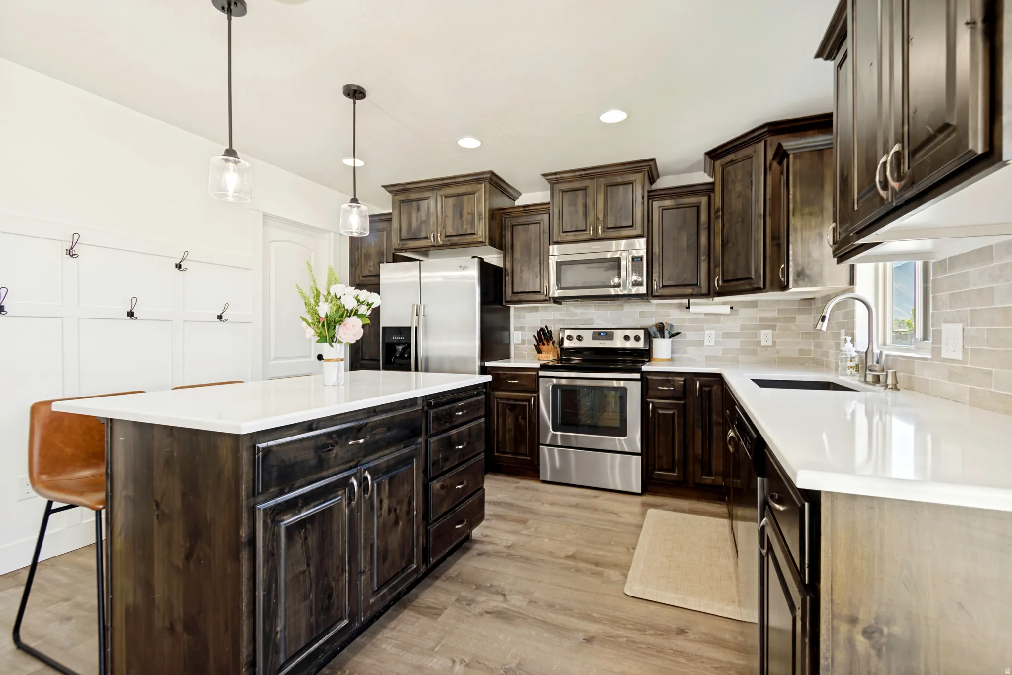 Kitchen with wood-finish flooring, dark wood cabinetry, light-toned countertops, stainless steel appliances, and a tiled backsplash.